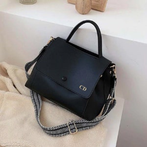 May include: Black leather handbag with a top handle and a long adjustable strap. The strap is black and white striped. The bag has a flap closure with a gold button and the initials "CD" are embossed on the flap.