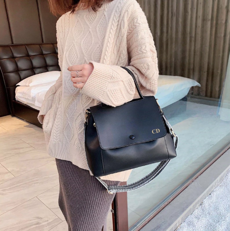 May include: Black leather satchel bag with a top flap and a gold buckle. The bag has a long adjustable strap and a shorter top handle. The initials "CD" are embossed on the front flap.
