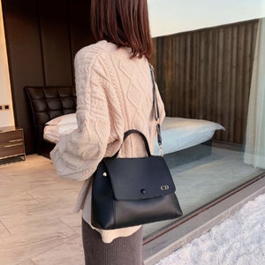 May include: A black leather satchel handbag with a top flap and a gold buckle closure. The bag has a short handle and a long adjustable strap. The initials "CD" are embossed on the front flap.