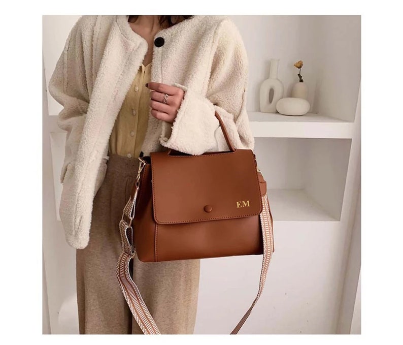 May include: A brown leather handbag with a flap closure and a decorative strap. The bag has a top handle and a detachable shoulder strap with a patterned design. The initials "EM" are visible on the front of the bag.