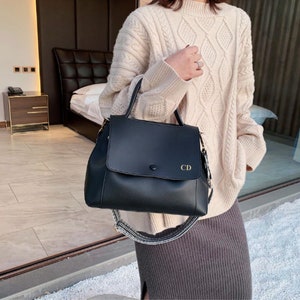 May include: A black leather satchel handbag with a gold buckle and a long strap. The bag has a flap with the initials "CD" on it.