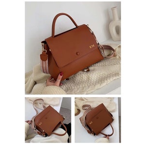 May include: A brown handbag with a top handle and a detachable shoulder strap. The bag features a flap closure and the initials "EM" in gold. The strap has a woven pattern in shades of brown and cream.