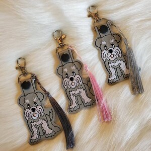 Puede incluir: Tres llaveros de cuero gris con dise&ntilde;os de perros schnauzer bordados. Cada llavero tiene un borla de un color diferente: rosa, negro y gris.