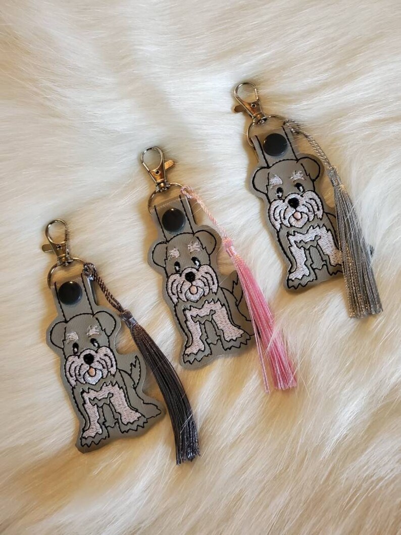 Puede incluir: Tres llaveros de cuero gris con dise&ntilde;os de perros schnauzer bordados. Cada llavero tiene un borla de un color diferente: rosa, negro y gris.