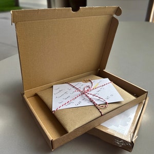 May include: A brown cardboard box with the lid open, revealing a brown paper package tied with red and white twine. A white card with handwritten text is visible on top of the package. The text reads "Dear Amanda, I am so happy for you.  Sending you lots of love.  Joy"