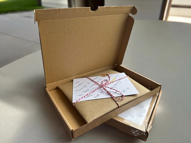 May include: A brown cardboard box with the lid open, revealing a brown paper package tied with red and white twine. A white card with handwritten text is visible on top of the package. The text reads "Dear Amanda, I am so happy for you.  Sending you lots of love.  Joy"