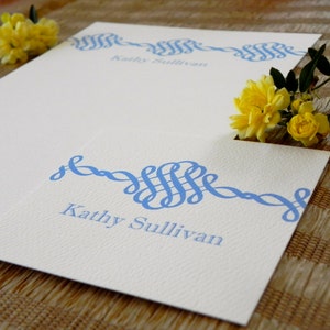 Peut inclure: Une carte blanche avec un motif d&eacute;coratif bleu et le nom "Kathy Sullivan" imprim&eacute; dessus. La carte est sur une surface en osier brune.