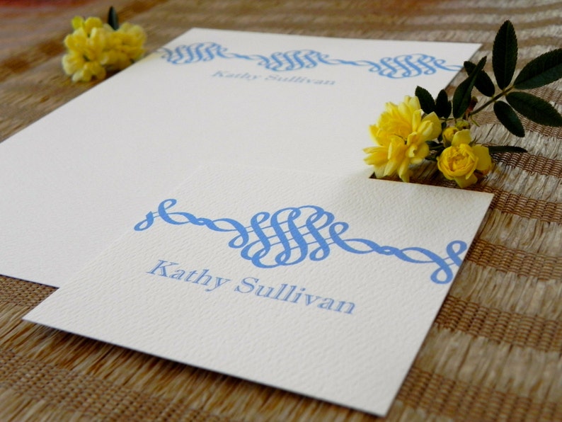 Peut inclure: Une carte blanche avec un motif d&eacute;coratif bleu et le nom "Kathy Sullivan" imprim&eacute; dessus. La carte est sur une surface en osier brune.