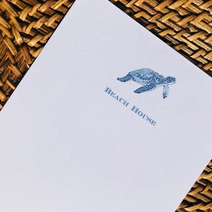Può includere: Un biglietto da visita bianco con un'illustrazione di tartaruga marina blu e il testo "Beach House" stampato in blu.