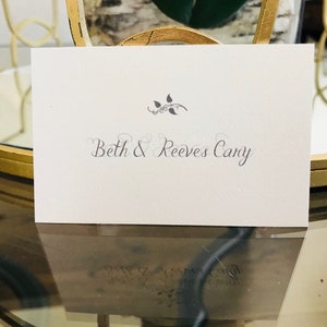 May include: A white place card with the names "Beth & Reeves Cary" in elegant script. A small floral design is above the names. The card is displayed on a reflective surface, suggesting a wedding or event setting.