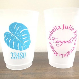 May include: Two white plastic cups with different designs. One cup has a blue monstera leaf design and the number 33480 printed on it. The other cup has a pink circle with the text "Virginia Emma Julie Isabella Megan Congrats!" printed on it.
