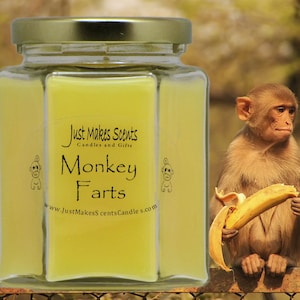 May include: A yellow glass jar candle with a gold lid. The label reads "Just Makes Scents Candles and Gifts" and "Monkey Farts" with a cartoon monkey on either side. The website address is "www.JustMakesScentsCandle.com".