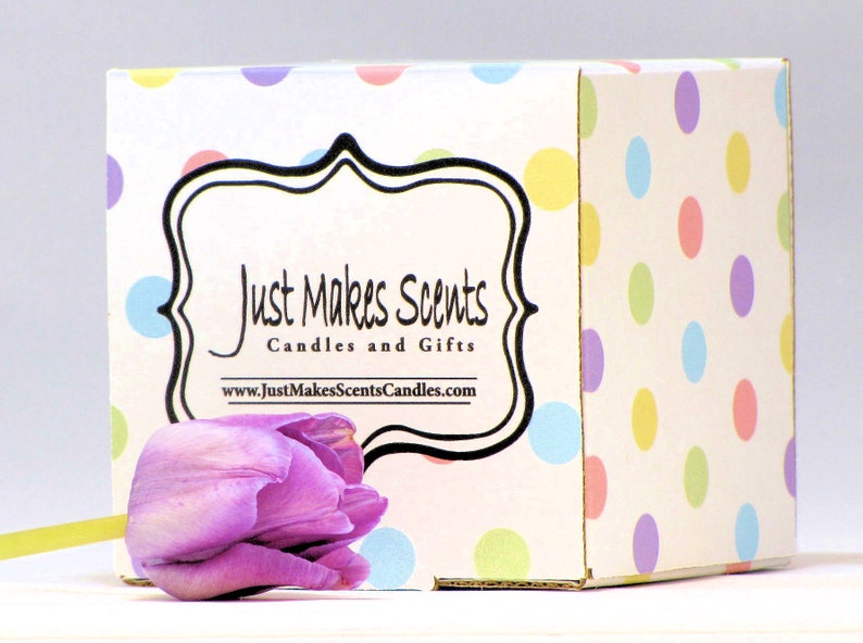 Op de afbeelding: Een witte doos met een stippenpatroon in pastelkleuren. De doos heeft een zwart label met de tekst "Just Makes Scents Candles and Gifts" en het websiteadres "www.JustMakesScentsCandles.com". Een paarse tulp bevindt zich op de voorgrond.