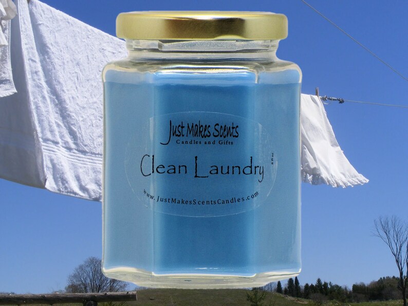 以下が含まれることがあります： 青いワックスと金の蓋が付いたガラス製のキャンドルが入った瓶。ラベルには「Just Makes Scents Candles and Gifts」と「Clean Laundry」とウェブサイトのアドレス「www.JustMakesScentsCandles.com」が書かれています。