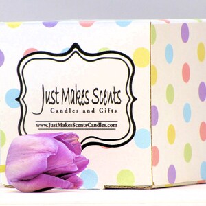 以下が含まれることがあります： パステルカラーのドット柄の白い箱。箱には、「Just Makes Scents Candles and Gifts」というテキストとウェブサイトアドレス「www.JustMakesScentsCandles.com」が書かれた黒いラベルが付いています。手前に紫色のチューリップがあります。