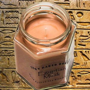 Op de afbeelding: Een zeshoekige glazen pot met een bruine sojawas kaars erin. De kaars is gelabeld met "Egyptian Musk" en "Hand Poured Soy Blend". De pot staat op een achtergrond van bruine en gouden hi&euml;rogliefen.