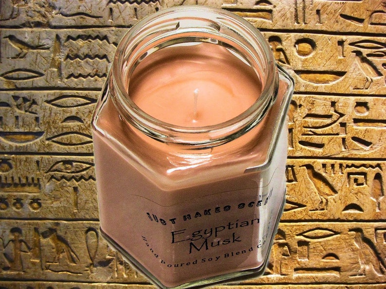Op de afbeelding: Een zeshoekige glazen pot met een bruine sojawas kaars erin. De kaars is gelabeld met "Egyptian Musk" en "Hand Poured Soy Blend". De pot staat op een achtergrond van bruine en gouden hi&euml;rogliefen.