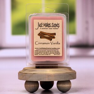 May include: A small, pink wax melt in a clear plastic container with a label that reads "Just Makes Scents Candles and Gifts" and "Cinnamon Vanilla". The wax melt is on a wooden stand with three round legs.