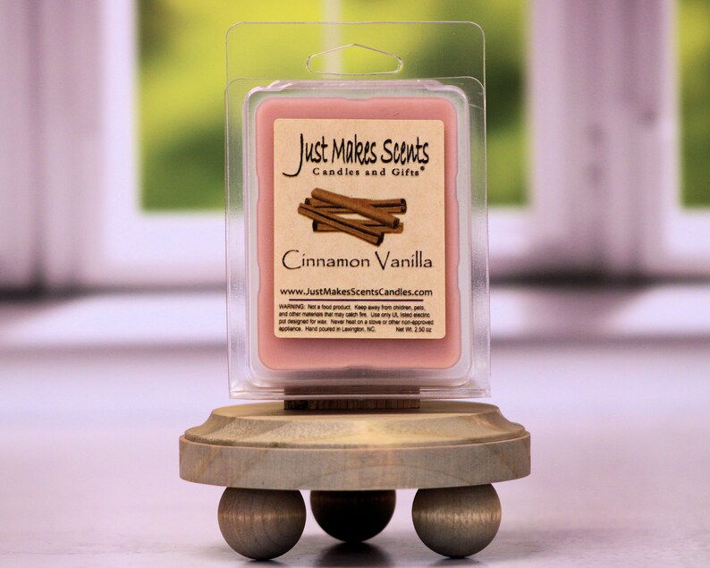 May include: A small, pink wax melt in a clear plastic container with a label that reads "Just Makes Scents Candles and Gifts" and "Cinnamon Vanilla". The wax melt is on a wooden stand with three round legs.