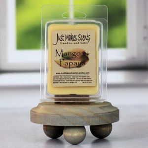 May include: A small, clear plastic container of wax melts with a label that reads "Just Makes Scents Candles and Gifts" and "Mango & Papaya". The container is sitting on a wooden stand with three legs.