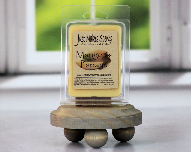 May include: A small, clear plastic container of wax melts with a label that reads "Just Makes Scents Candles and Gifts" and "Mango & Papaya". The container is sitting on a wooden stand with three legs.