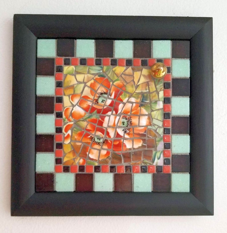 May include: Square mosaic art piece with a black frame. The mosaic features a central image of orange poppies, surrounded by a border of brown, mint green, and red tiles. A small, round, gold-colored embellishment is in the upper right corner.