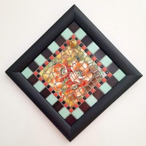May include: A mosaic artwork featuring orange and red poppy flowers. The artwork is framed in a black square frame with a decorative border of alternating teal and brown squares. A gold-colored glass bead is in the center.