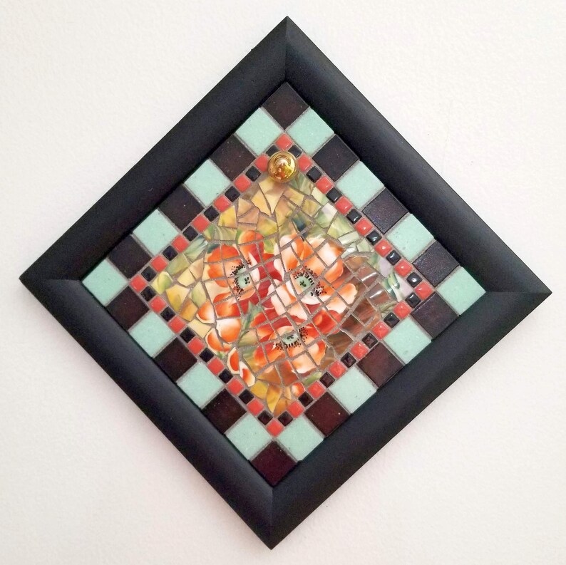 May include: A mosaic artwork featuring orange and red poppy flowers. The artwork is framed in a black square frame with a decorative border of alternating teal and brown squares. A gold-colored glass bead is in the center.