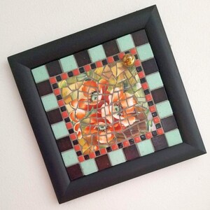 May include: Square mosaic art piece with a black frame. The mosaic features a floral design with red, orange, and yellow flowers. The border is a checkerboard pattern of brown, mint green, and red tiles. A small, gold-colored glass marble is affixed to the top.