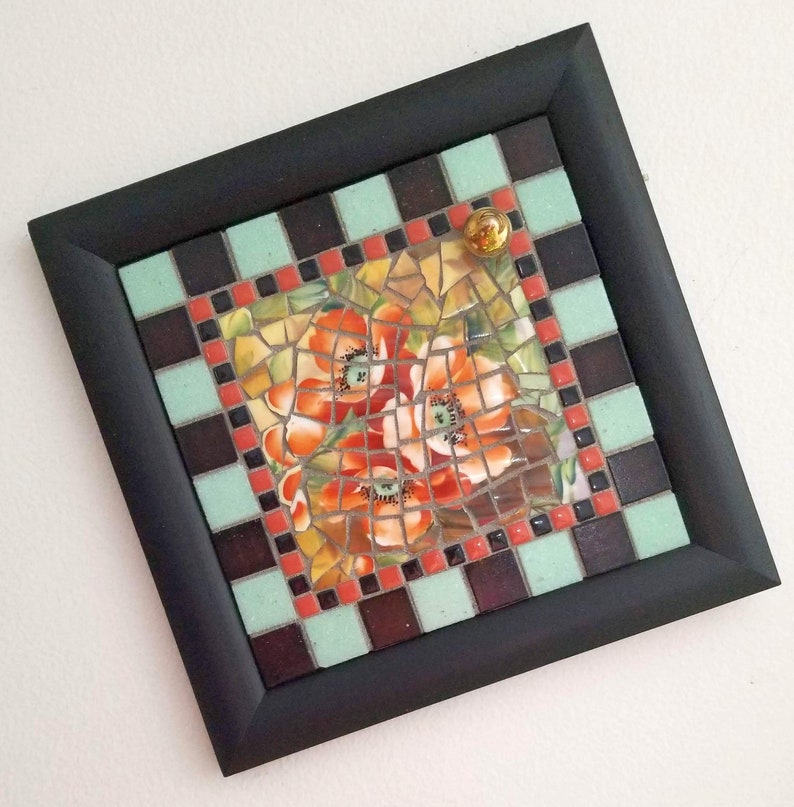 May include: Square mosaic art piece with a black frame. The mosaic features a floral design with red, orange, and yellow flowers. The border is a checkerboard pattern of brown, mint green, and red tiles. A small, gold-colored glass marble is affixed to the top.