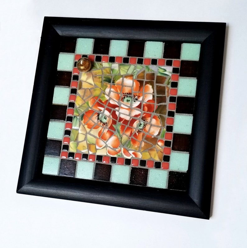 May include: Square mosaic art piece with a black frame. The mosaic features a floral design with orange and red flowers. The border has a checkerboard pattern of black, brown, and light green tiles. A small, round, gold-colored embellishment is in the upper left corner.