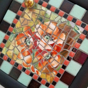 May include: A square mosaic tile art piece with a black frame. The mosaic features orange and yellow flowers with green centers. The border includes black, red, and light green tiles, and a gold knob.