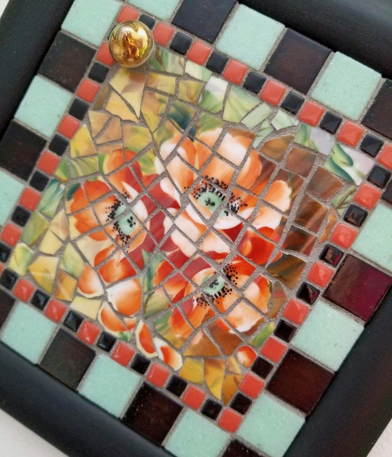 May include: A square mosaic tile art piece with a black frame. The mosaic features orange and yellow flowers with green centers. The border includes black, red, and light green tiles, and a gold knob.