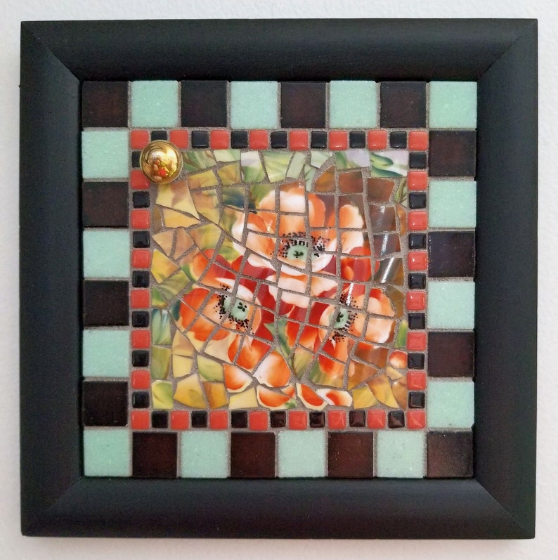 May include: Square mosaic art piece with a black frame. The mosaic features orange flowers with green leaves. The border includes a checkerboard pattern of brown, mint green, and red tiles. A gold-colored knob is in the upper left corner.