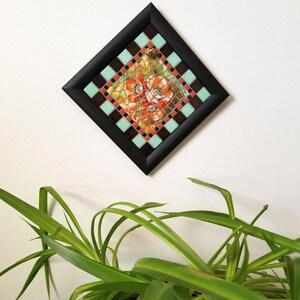 May include: A framed mosaic tile art piece. The square frame is black, surrounding a diamond-shaped mosaic. The mosaic features a floral design with red, orange, and yellow flowers. The mosaic is bordered by alternating squares of brown, teal, and red.