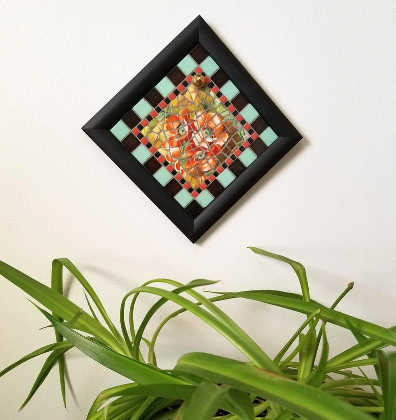 May include: A framed mosaic tile art piece. The square frame is black, surrounding a diamond-shaped mosaic. The mosaic features a floral design with red, orange, and yellow flowers. The mosaic is bordered by alternating squares of brown, teal, and red.