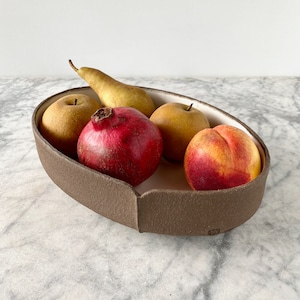 Könnte beinhalten: Eine braune Keramikschüssel mit weißem Innenraum, gefüllt mit Obst. Die Schüssel ist oval geformt und hat einen leicht erhöhten Rand. Das Obst umfasst einen roten Granatapfel, eine gelbe Birne und zwei gelbe Äpfel. Die Schüssel steht auf einer weißen Marmorfläche.