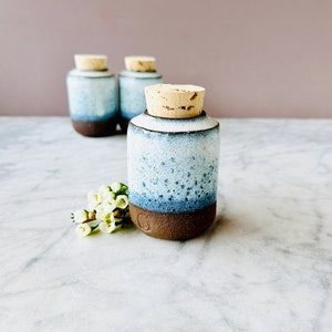 May include: Three small ceramic jars with cork lids. The jars are glazed in a light blue and white colour with a dark brown base. The jars are arranged in a row on a white marble surface.