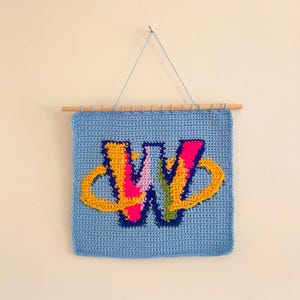 May include: A handmade, square, light blue crochet wall hanging featuring a colorful letter "W". The letter is primarily pink, yellow, and orange, with a blue outline. A wooden dowel and string are used for hanging.