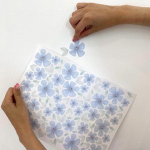 May include: A sheet of light blue floral wall decals with a matching flower being applied to a white wall. The decals feature a watercolor design of blue flowers and green leaves, perfect for home decor.