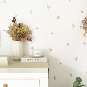 Könnte beinhalten: Eine weiße Wand mit einem hellbraunen Blumenmuster. Eine weiße Vase mit getrockneten Blumen steht auf einer weißen Kommode mit einer Kerze und einem Buch mit dem Titel "Matisse" darauf.