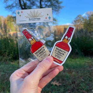 May include: Two Maker's Mark Bourbon Whiskey stickers. One sticker is loose, and the other is in a clear plastic bag with a Fernweh Haus tag. The stickers are red, white, and tan, with the brand name and product information.