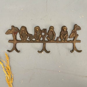 May include: A bronze-colored wall hook featuring five monkey figures in the "See no evil, hear no evil, speak no evil" pose. The monkeys are perched on a branch with three hooks below for hanging items. The hook is mounted on a gray surface.