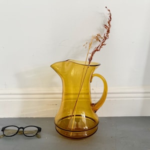 May include: Amber glass pitcher with a wide mouth and a handle. The pitcher is empty and has a dried flower stem inside.