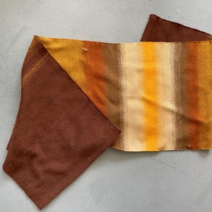 May include: A woven scarf with a color gradient of brown, orange, and yellow. The scarf is folded, showing a solid brown section and a striped section with vertical bands of color. The fabric appears to be textured.