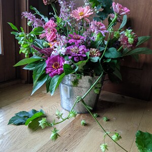 Puede incluir: Un ramo de flores rosas y moradas en un cubo de metal galvanizado. Las flores est&aacute;n dispuestas con hojas verdes y una vid con brotes verdes.