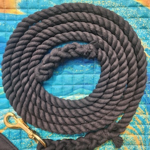 May include: A thick, black cotton rope lead coiled in a circular shape. The rope features a braided section near the end and is attached to a polished brass swivel snap hook. The texture of the rope is twisted and sturdy, designed for equine use.