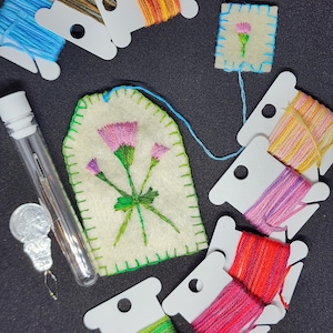 May include: An assortment of embroidery supplies on a dark surface. The image features spools of colorful thread, a needle case, and two embroidered felt pieces with floral designs. The felt pieces are stitched with pink and green thread, and the spools contain various colors.