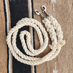 May include: Cream-colored rope leash with two silver-colored metal clasps. The leash is coiled and braided, with a tassel at the end. The background is a tan, black, and white patterned textile.