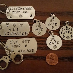 May include: A set of seven silver keychains with various firefighter-themed designs. The keychains feature phrases like "Fire Fighters Kick Ash", "911 Dispatch", "I've Got Your 6 Always", "Be Safe", and "Smoke Eater".
